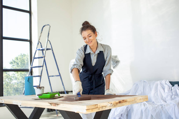 молодая темноволосая женщина в перчатках улыбается, готовая работать в мастерской по покраске поверхности стола роликом - Фото, изображение