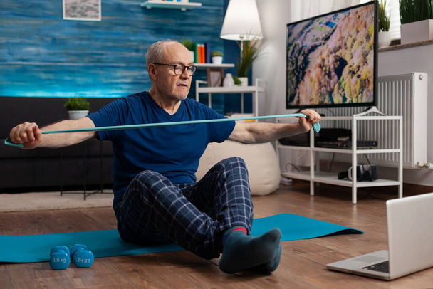 Pensionato che lavora alla resistenza del corpo che esercita i muscoli delle braccia con elastico seduto su tappetino yoga - Foto, immagini