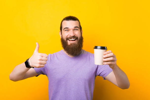 Joven sonriente barbudo bebiendo sostener papel taza de café mostrar el pulgar hacia arriba gesto aislado sobre fondo amarillo - Foto, Imagen