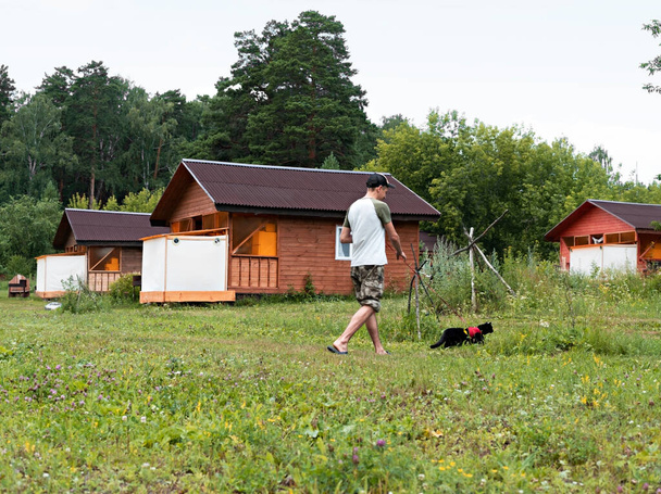 young man in cap walking with black cat in red harness on leash next to wooden houses on green grass, friendship pet love and care, outdoor - Fotoğraf, Görsel