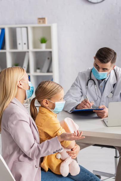 Woman hugging daughter and talking to doctor in medical mask writing on clipboard  - Fotoğraf, Görsel