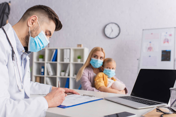 Doctor en escritura máscara médica en portapapeles cerca de los dispositivos y la familia en la clínica  - Foto, imagen