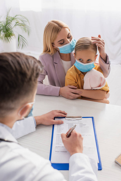 Mother in protective mask touching child with toy near family doctor writing prescription - Foto, Imagen