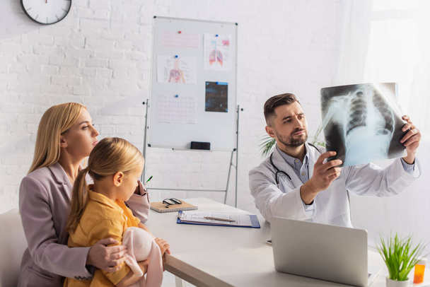 Pediatrician looking at fluorography near family and laptop in clinic  - Foto, Imagen
