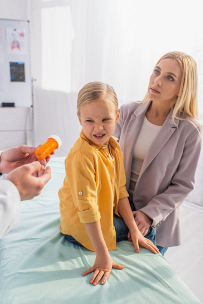 Gyermekorvos kezében tabletták közelében undorodott gyerek orvosi kanapén és anya  - Fotó, kép