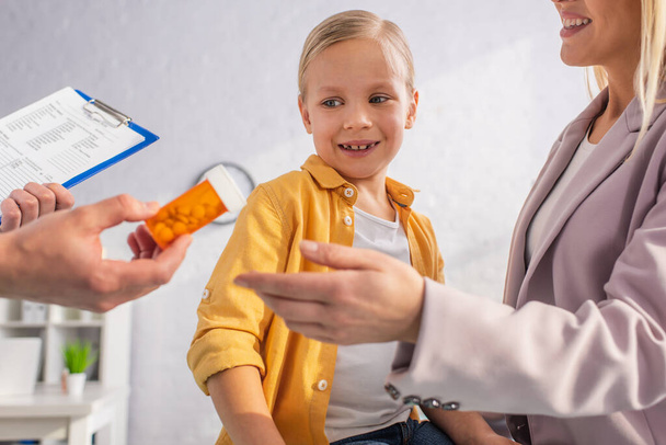 Médecin avec presse-papiers tenant des pilules près de la mère et de l'enfant souriants  - Photo, image