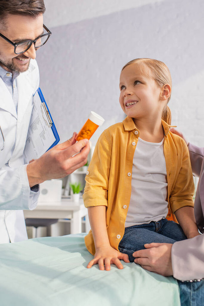 Positieve arts met klembord houden pillen in de buurt glimlachend meisje op de medische bank en vrouw  - Foto, afbeelding