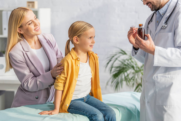 Pediatrician holding syrup near smiling mother and child on medical couch  - Photo, Image