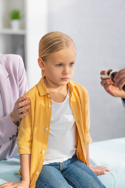 Kid looking at blurred family doctor with syrup near mother in hospital  - Photo, Image