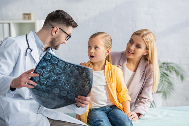Pediatrician holding blurred mri scan near amazed kid and mother - Foto, Imagen