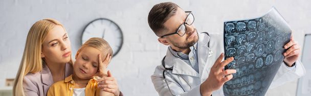 Pediatrician in eyeglasses looking at mri scan near mother and sick child, banner  - Фото, изображение