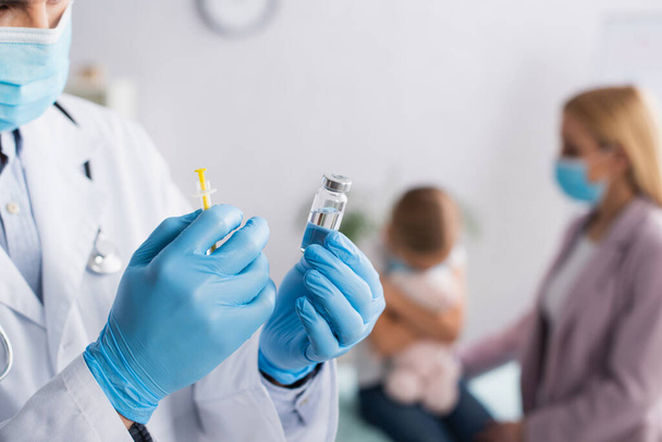 Лікар у медичній масці, що тримає вакцину біля розмитої матері та дитини
  - Фото, зображення