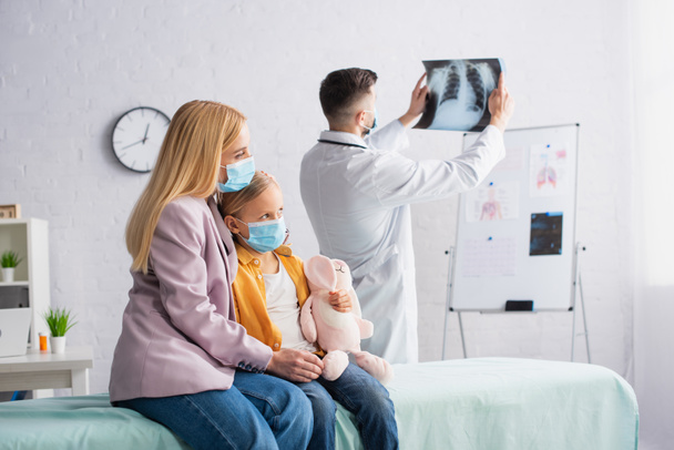 Γυναίκα με ιατρική μάσκα αγκαλιάζει το παιδί στον ιατρικό καναπέ κοντά στο γιατρό με φθοριογραφία στην κλινική  - Φωτογραφία, εικόνα