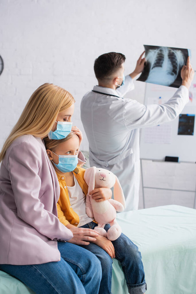 woman in medical mask sitting near kid on medical couch and blurred pediatrician with fluorography - Photo, image