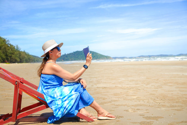 Femme asiatique élégante portant une longue robe bleue. Bain de soleil au bord de la mer tenant un smartphone pour prendre des photos de beaux paysages naturels Voyage à la mer en Thaïlande, tourisme - Photo, image