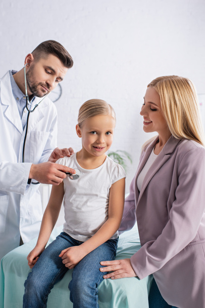 Lächelndes Kind blickt mit Stethoskop in die Kamera neben Mutter und Arzt  - Foto, Bild