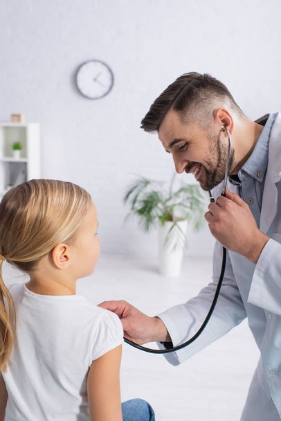 Cheerful pediatrician examining kid with stethoscope in hospital  - Photo, Image