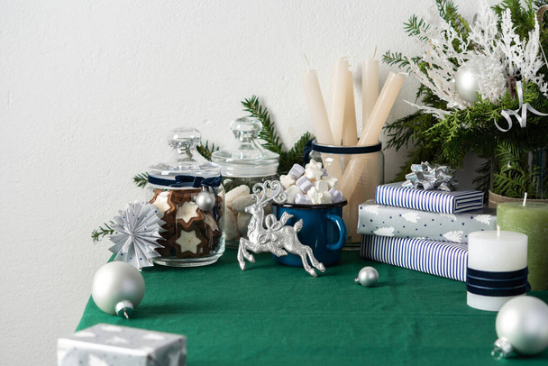 Festlich geschmückter Weihnachts- oder Neujahrstisch, Thuja und dekorative Fichtenzweige, Kerzen und Weihnachtskugeln, Plätzchen im Glas und ein blauer Eisenkrug mit Marshmallows, Geschenkschachteln und Silberhirschen - Foto, Bild