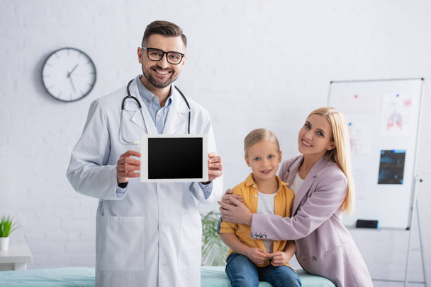 Positivo medico di famiglia in possesso di tablet digitale vicino madre abbracciando ragazza sul divano medico  - Foto, immagini