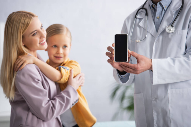 Педиатр держит смартфон с чистым экраном рядом с матерью, обнимающей дочь в клинике  - Фото, изображение