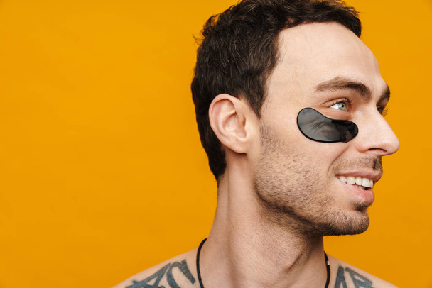 Close up of a shirtless mid aged brunette man wearing cosmetic eye patches over yellow wall background - Foto, imagen