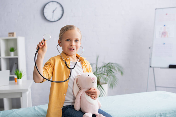 Excited child in stethoscope holding soft toy on medical couch in hospital  - Foto, Bild