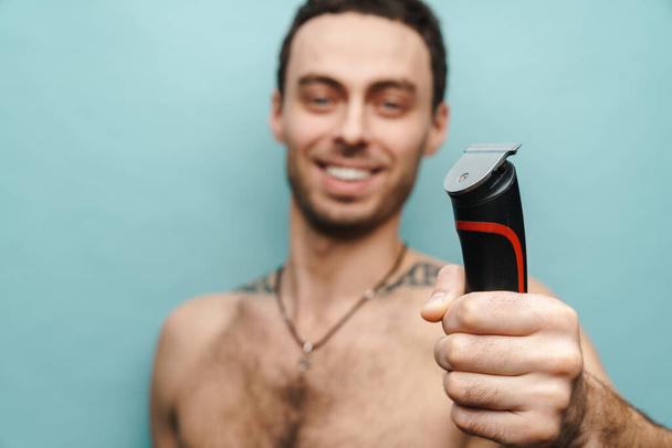 Close up portrait of a smiling middle aged shirtless man holding electric shaving over blue wall background - 写真・画像