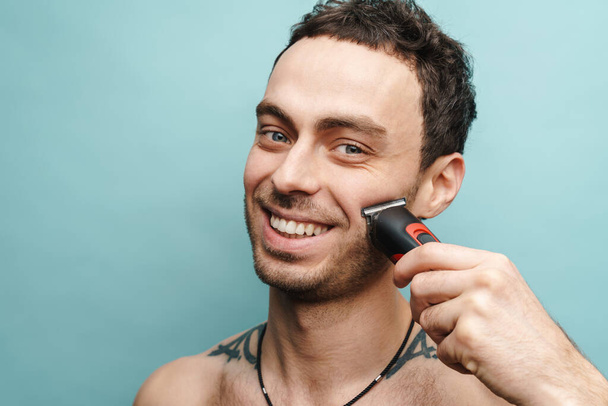 Close up portrait of a smiling middle aged shirtless man holding electric shaving over blue wall background - Zdjęcie, obraz