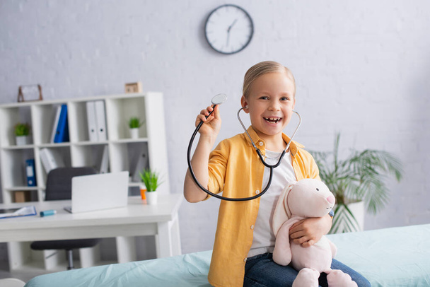 Happy girl with stethoscope and soft toy looking at camera in hospital  - Foto, imagen