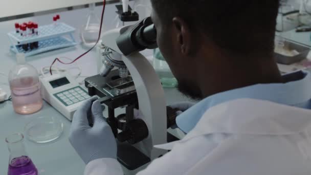 Handheld slowmo over shoulder shot of black male scientist in white coat and gloves looking into microscope while working in lab - Séquence, vidéo