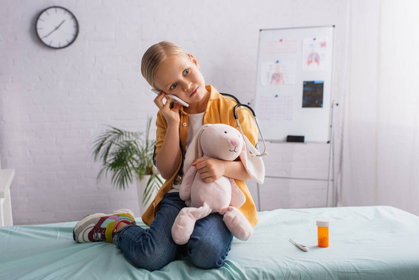criança com coelho de brinquedo sentado no sofá médico e falando no smartphone - Foto, Imagem