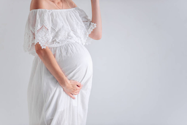 Close-up A young pregnant woman in anticipation of a baby in a white hat, dress, hugs her belly. Tender girl in a minimalistic, isolated background copy space. Happy maternity pregnancy, surrogacy - Photo, image
