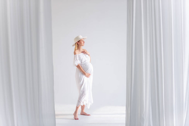 Young pregnant woman in anticipation of a baby in a white hat, dress, hugs her belly. Tender blonde girl in a minimalistic, isolated interior of airy fabrics. Happy maternity pregnancy, surrogacy. - Фото, изображение