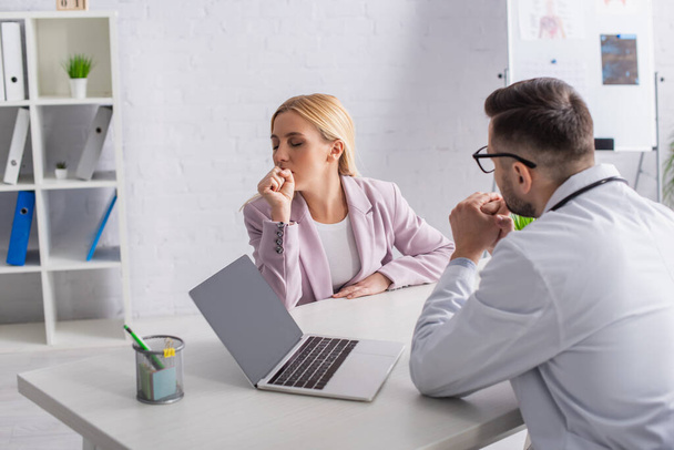 médico sentado cerca de la computadora portátil con pantalla en blanco mientras mira a la mujer tosiendo durante la consulta - Foto, imagen