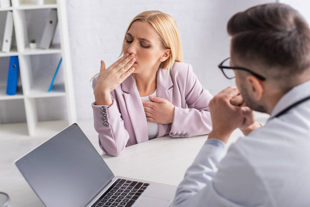 woman coughing near blurred physician and laptop with blank screen in consulting room - Photo, Image