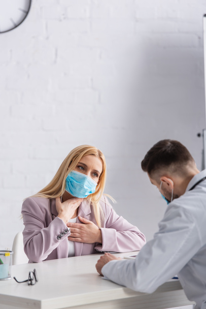 Kranke Frau in medizinischer Maske berührt Brust und Rachen in der Nähe von Arzt - Foto, Bild