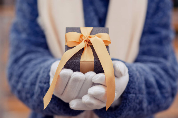 Woman dressed in a blue winter coat holding a gift box with a silk ribbon at the street - Photo, Image