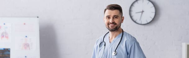 happy physician wearing blue uniform and stethoscope on neck looking at camera in clinic, banner - Foto, Imagen