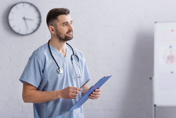 smiling doctor in blue uniform looking away while holding clipboard - Фото, изображение