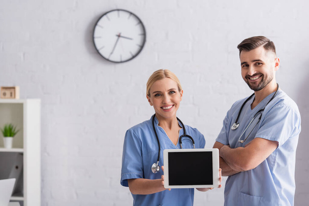 veselá zdravotní sestra ukazující digitální tablet s prázdnou obrazovkou poblíž pozitivního lékaře stojící se zkříženýma rukama - Fotografie, Obrázek