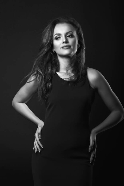 Fashion studio portrait of a pretty brunette woman with perfect makeup and hairstyle wears classic black dress posing in a dark studio. Black and white shot - Фото, изображение