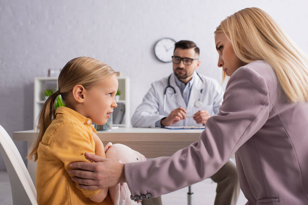 ustaraný dítě při pohledu na maminku v blízkosti rozmazané lékaře v nemocnici - Fotografie, Obrázek
