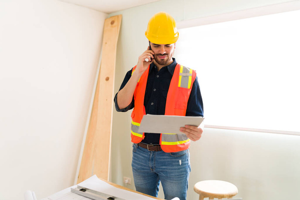 Glückliche männliche Arbeiter und Auftragnehmer überprüfen die Baupläne, während sie mit dem Kunden oder Lieferanten telefonieren - Foto, Bild