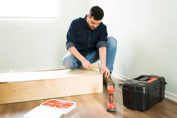 Attractive young man and handyman using a hammer and building a new wooden bookshelf for his home   - Photo, Image
