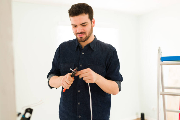 Χαρούμενος Λατίνος τεχνίτης που κόβει με τσιμπιδάκι τα ηλεκτρικά καλώδια για να εγκαταστήσει ένα νέο διακόπτη στο σπίτι του - Φωτογραφία, εικόνα