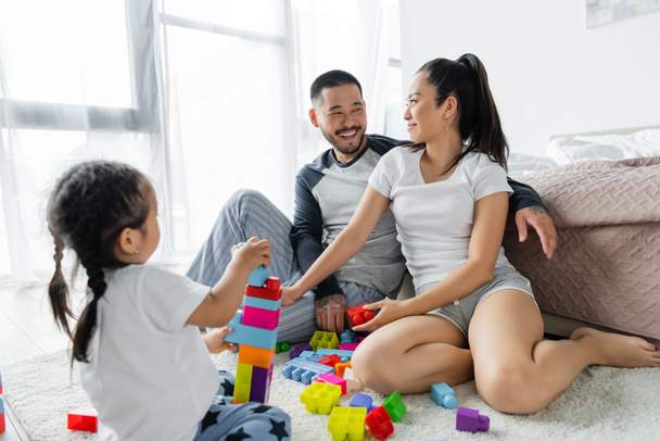 happy asian husband and wife looking at each other while toddler daughter playing building blocks  - Zdjęcie, obraz