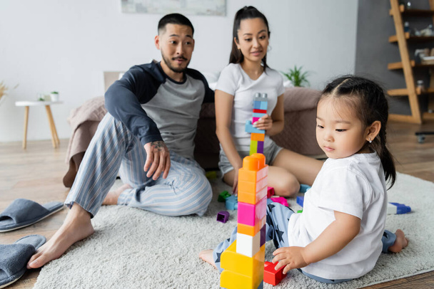 blurred asian husband and wife looking at toddler daughter playing building blocks  - Valokuva, kuva
