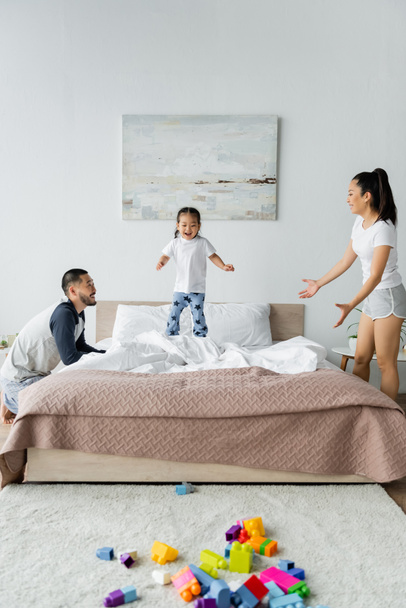 happy asian parents looking at toddler daughter jumping on bed - Фото, зображення