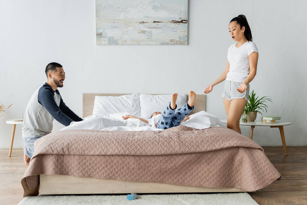 happy asian husband and wife looking at toddler daughter jumping on bed - Zdjęcie, obraz