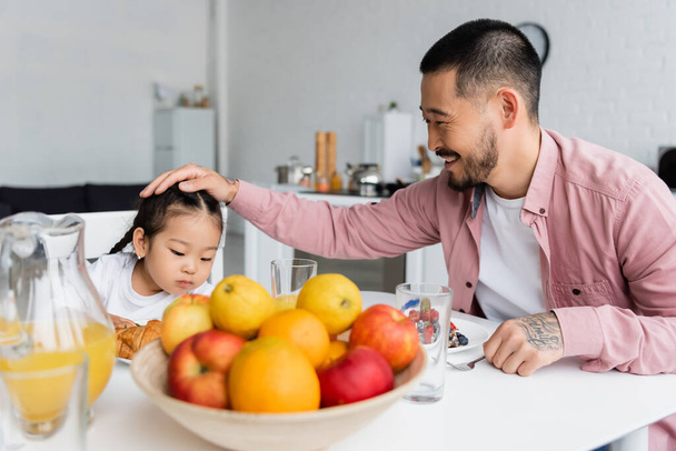 happy father stroking hair of asian daughter during breakfast  - Фото, изображение